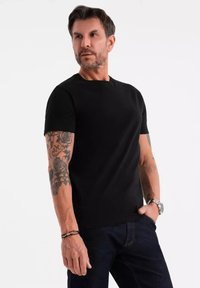 Černé tričko s krátkým rukávem vyrobené z měkké tkaniny, s kulatým výstřihem a přiléhavým střihem. Ozdobené ruce tetováním, nošeno s tmavými denimovými džíny.