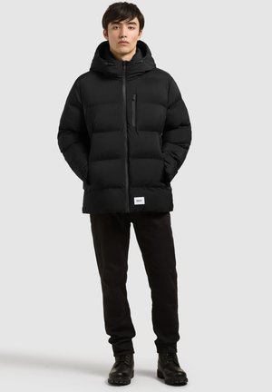 Jeune homme debout portant une doudoune noire à capuche, un pantalon noir et des bottes noires sur un fond gris clair uni.