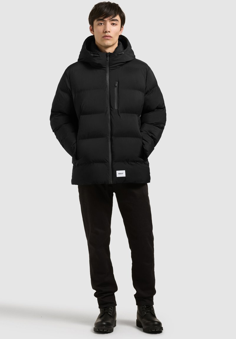 Jeune homme debout portant une doudoune noire à capuche, un pantalon noir et des bottes noires sur un fond gris clair uni.