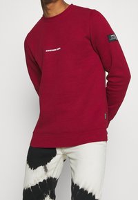 Sweat-shirt à col rond rouge avec le texte "REDEFINED REBEL" en blanc sur la poitrine et un écusson sur la manche.