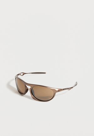 Lunettes de soleil enveloppantes bronze avec des verres ovales teintés et des branches fines et courbées reposant sur une surface blanche.