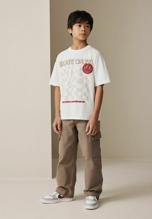 Boy standing wearing white "SKATE DAZE" graphic T-shirt, beige cargo pants, and white sneakers against plain beige background.