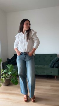 Žena v zdobenej bielej košeli, širokých džínsach a hnedých sandáloch stojí s rukami v vreckách pri zelenej pohovke a kvetináči v jasnej miestnosti.