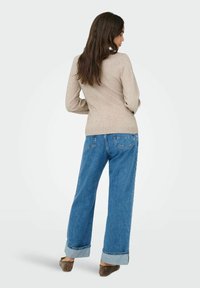 Un pull beige au design ajusté, associé à un jean en denim bleu ample avec des ourlets retroussés. Le look est complété par des chaussures à enfiler marron.