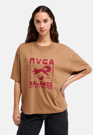 Młoda kobieta z długimi ciemnymi włosami, ubrana w luźny brązowy t-shirt z czerwonym napisem i wzorem „RVCA Balance of Opposites”, w połączeniu z czarnymi dżinsami.