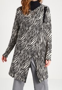 Zebramönstrad stickad lång kappa i svart och kräm, med en sidoklippning och svart accentrem på ena axeln. Mjuk textur.