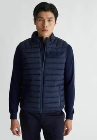 Gilet imbottito blu navy con collo alto, design trapuntato, tasche con zip e una superficie liscia. Presenta dettagli con logo sul petto.