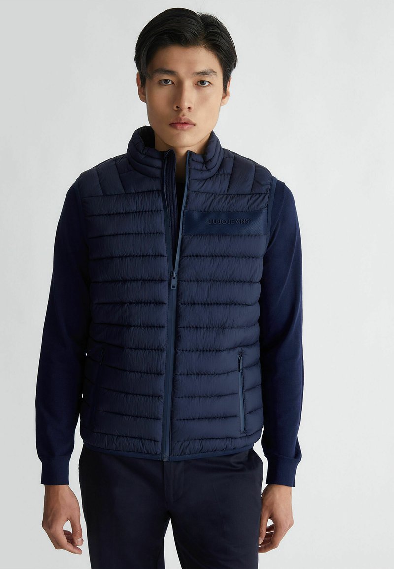Gilet imbottito blu navy con collo alto, design trapuntato, tasche con zip e una superficie liscia. Presenta dettagli con logo sul petto.