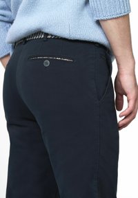 Navyblauwe broek met een gladde textuur, voorzien van een decoratieve zak met een metallic accent en zijzakken. Gedragen met een lichtblauwe trui.