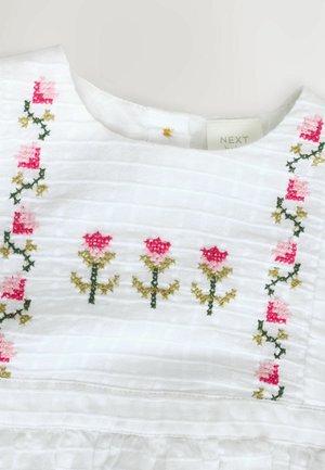 Valkoinen kankainen mekko, jossa on rakenneraitoja. Kauluksessa ja vartalolla on kukkakirjontaa vaaleanpunaisena ja vihreänä. Konepesuohje näkyvissä.