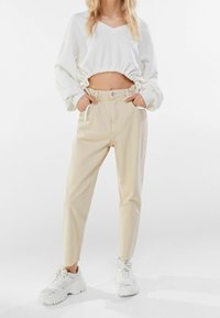 Kvinna som bär beige höga midjebyxor, vit kortärmad topp med lång ärm och klumpiga vita sneakers, står med händerna i fickorna.