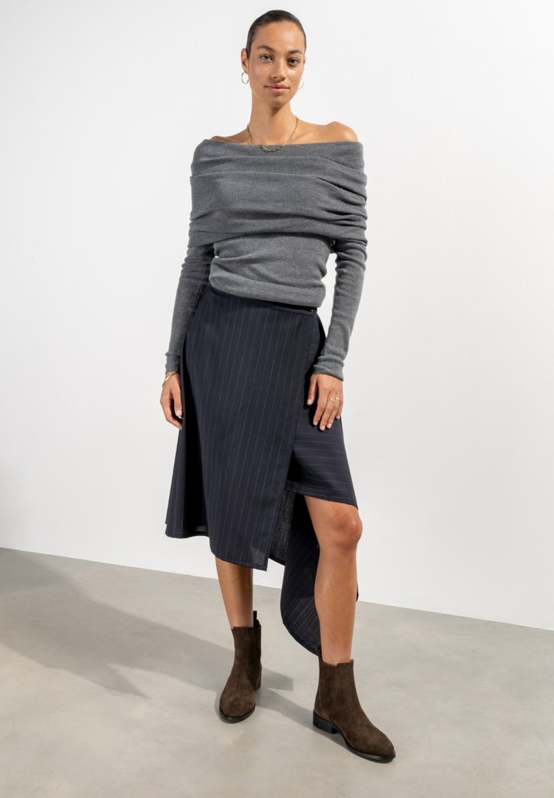 Un pull gris à épaules dénudées associé à une jupe asymétrique à rayures foncées. Des bottines marron complètent la tenue. Fonds uni.