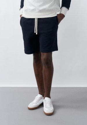 Személy fekete zsinóros rövidnadrágot, fehér, gumitalpú tornacipőt és fehér-kék hosszú ujjú felsőt visel, és zsebébe tett kézzel áll.