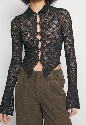 Mujer con un top negro de encaje ajustado de manga larga con cierres de botones en forma de ojo de cerradura y pantalones marrones de pana de cintura alta.