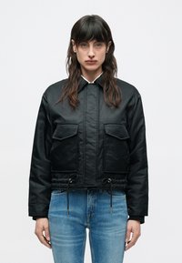 Μαύρο cropped μπουφάν με δύο εμπρόσθιες τσέπες, κορδόνι στο τελείωμα και λείο υλικό. Συνδυασμένο με εφαρμοστά ανοιχτό μπλε τζιν.