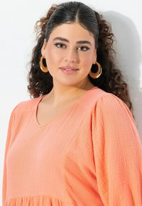 Mujer con cabello oscuro y rizado que lleva pendientes de aro dorados grandes y una blusa de melocotón texturizada, mirando suavemente a la cámara contra un fondo blanco.