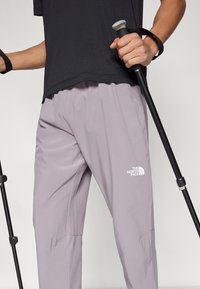 Ανοιχτό μοβ ελαφριά παντελόνια με ελαστική μέση και λογότυπο, συνδυασμένα με μαύρο πουκάμισο και δύο μπαστούνια πεζοπορίας. Λείο ύφασμα.