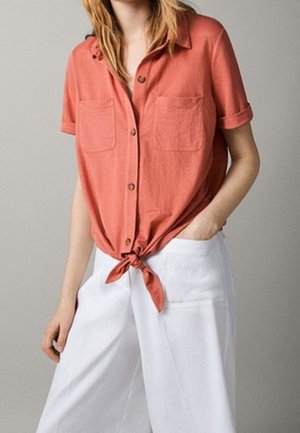 Koraalkleurig overhemd met korte mouwen, knoopsluiting, twee borstzakken en een strik aan de taille, gecombineerd met witte wijde broek.