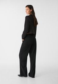 Svart kort blazer med långa ärmar, kombinerad med lösa svarta byxor. Båda plaggen har en mjuk tygkvalitet och en minimalistisk design.