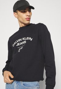 Felpa nera con testo bianco "CALVIN KLEIN JEANS", scollo tondo, polsini e orlo a costine; abbinata a jeans azzurri.