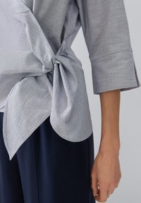 Blusa grigia con texture, caratterizzata da un nodo laterale, maniche tre quarti e orlo asimmetrico, abbinata a pantaloni scuri.