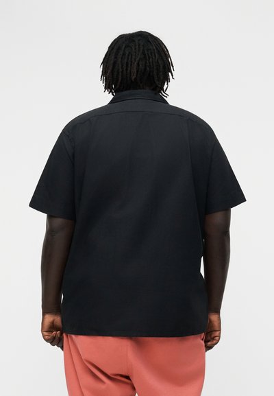 Osoba o ciemnej karnacji ze dreadlockami, ubraną w czarną koszulkę z krótkim rękawem i różowe spodnie, stojąca tyłem na jednolitym tle.