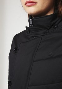 Svart puffjacka med hög krage, knapp- och dragkedjestängning samt en logotyp på bröstet. Slät, strukturerad tyg.