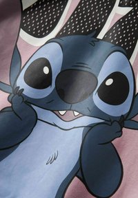 Postać z kreskówki Stitch, niebieska z dużymi oczami i nosem, uśmiechnięta i ciągnąca za policzki na różowym tle.