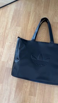 Juodas dirbtinės odos rankinė su dviem tvirtais rankenomis, centre iškilmingai pavaizduota Adidas logo. Lygus paviršius, stačiakampio formos.