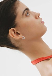 Pendiente de aro plateado con un diseño retorcido, que refleja la luz. Lleva puesto un modelo con el cabello oscuro recogido, mostrando el detalle de la oreja.