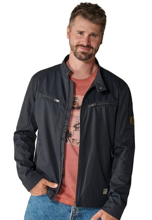 Dark navy bomber jacket made of lightweight material, featuring a stand collar, zippered pockets, and patches on the sleeves. Worn over a red shirt.
