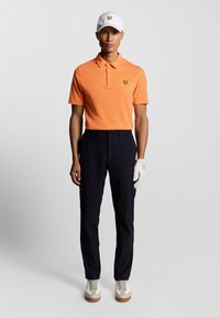 Polo skjorte i lys orange, parret med marineblå bukser. Hvid kasket med et logo; modellen bærer hvide sko med en struktureret sål og en golfhandsker.