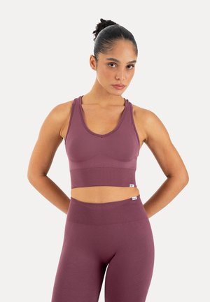 Kvinde iført matchende lilla sømløs sports-bh og højtaljede leggings, stående med hænderne bag ryggen mod en ensfarvet baggrund.