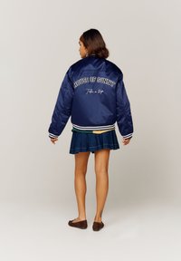 Veste bomber en satin bleu marine avec des poignets rayés blancs et verts, ornée de la broderie "HOUSE OF SUNNY" et "Take a trip" au dos.