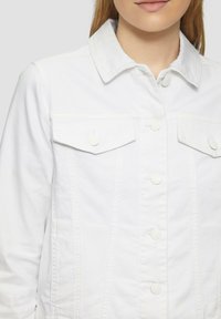 Weiße Jeansjacke mit spitzem Kragen, zwei Brusttaschen mit Klappen und Frontknopfverschluss mit passenden Knöpfen.