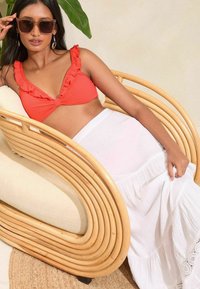Top bikini rosso a coste con bordi arricciati, abbinato a una gonna maxi bianca con tessuto texturizzato e dettagli in pizzo, su una sedia in rattan.