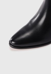 Botín negro de cuero hasta el tobillo con punta afilada, textura lisa y suela negra delgada. Presenta un diseño elegante y minimalista sin adornos.
