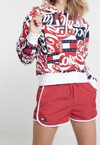 Sweat à capuche court rouge et blanc avec les logos Coca-Cola et Tommy Jeans, doté d'une capuche à cordon de serrage. Associé à un short rouge avec une bordure blanche.