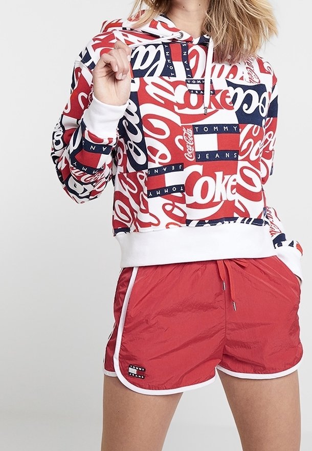 Sweat à capuche court rouge et blanc avec les logos Coca-Cola et Tommy Jeans, doté d'une capuche à cordon de serrage. Associé à un short rouge avec une bordure blanche.