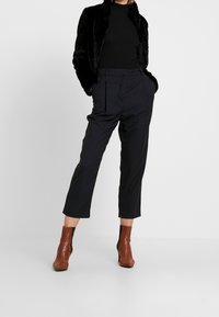 Giacca in ecopelliccia nera, dolcevita nera aderente, pantaloni a scacchi scuri cropped, stivaletti alla caviglia con tacco blocco marroni.