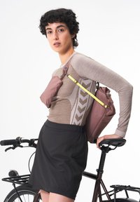 Braunes langärmliges Oberteil mit strukturiertem Muster, kombiniert mit einem schwarzen Rock. Rosa Tasche mit einem gelben Riemen gesichert, während ein Fahrrad in der Nähe steht.