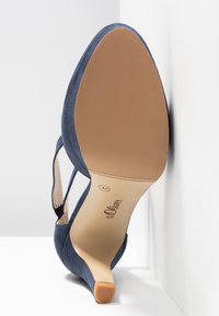 Chaussure à talon haut en daim bleu montrant une semelle beige lisse taille 37 avec le logo de la marque s.Oliver, inclinée contre un mur blanc d’angle.