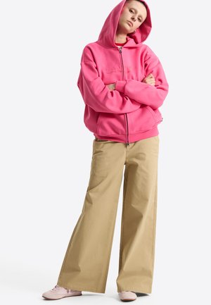 Person in a pink zip-up hoodie with the hood up, khaki wide-leg pants, and light pink shoes standing with arms crossed.