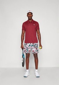 Crvena polo majica s ovratnikom, bijele kratke hlače s uzorkom u zelenim i ružičastim naglascima, bijela kapa, drži teniski reket i lopticu.