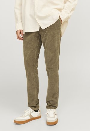 Olive green corduroy pants with a slim fit, featuring vertical ribbing, paired with white and tan sneakers. The shirt is light-colored and loose-fitting.