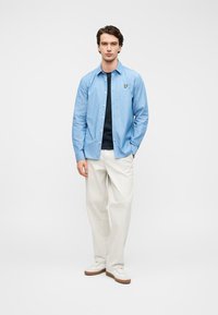 Γαλάζιο πουκάμισο με λωρίδες και κουμπιά, φορεμένο πάνω από σκούρα μπλούζα. Συνδυασμένο με άσπρο παντελόνι και άσπρα αθλητικά παπούτσια με καφέ λεπτομέρειες.