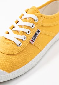 Sneaker in tela gialla con lacci bianchi, punta in gomma e suola testurizzata. Presenta un'etichetta con il logo di lato e dettagli cuciti sulla parte superiore.