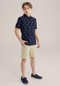 Mörkblå piké med vitt mönster, kort ärm, och två knappslå, parat med beige shorts och mörkblå slip-on skor.
