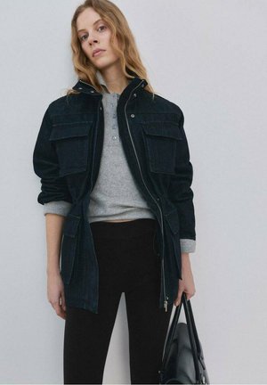 Jeune femme aux cheveux blonds ondulés portant une veste noire par-dessus une chemise grise boutonnée, un pantalon noir et tenant un sac à main noir.