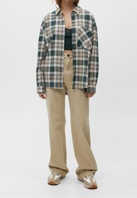 Vrouw in losse beige broek, groene cropped top, groen-beige geruit overhemd met knopen en witte sneakers staande tegen een witte achtergrond.